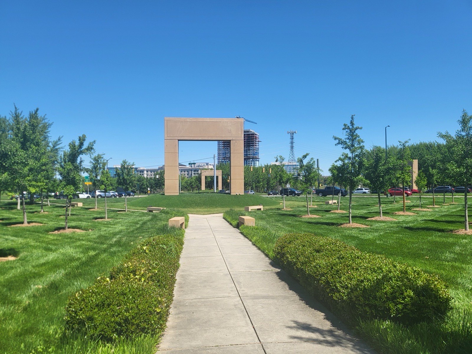 Tall stone arch sculpture on a manicured lawn with a walking path and young trees, framed against Ballantyne office towers in south Charlotte, North Carolina