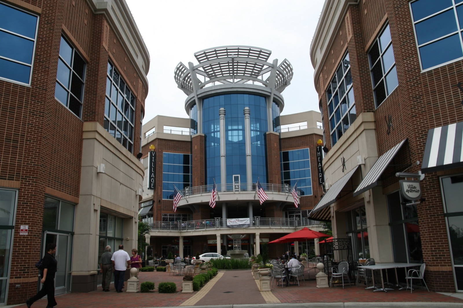 Brick and glass rotunda entrance of the Ballantyne Village complex with outdoor cafe seating, red umbrellas and American flags, south Charlotte