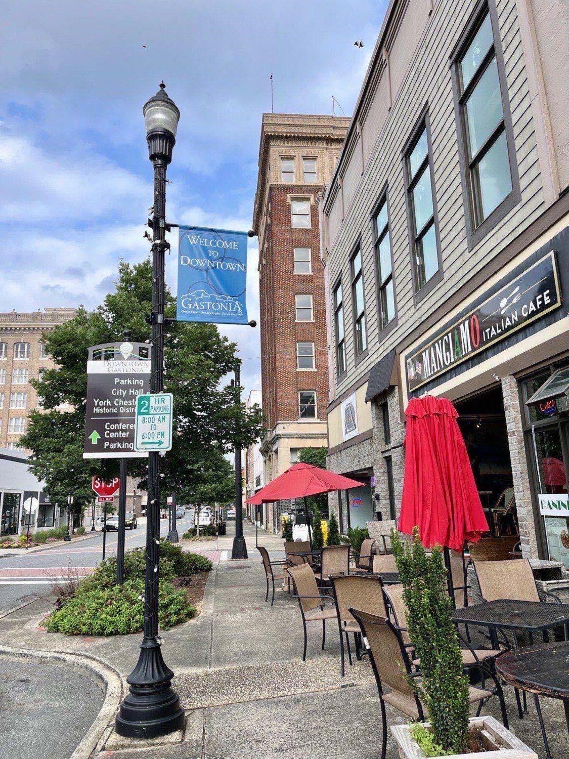 Street corner in downtown Gastonia, North Carolina with a Welcome to Downtown Gastonia pole banner, wayfinding signs, sidewalk cafe tables and red umbrellas