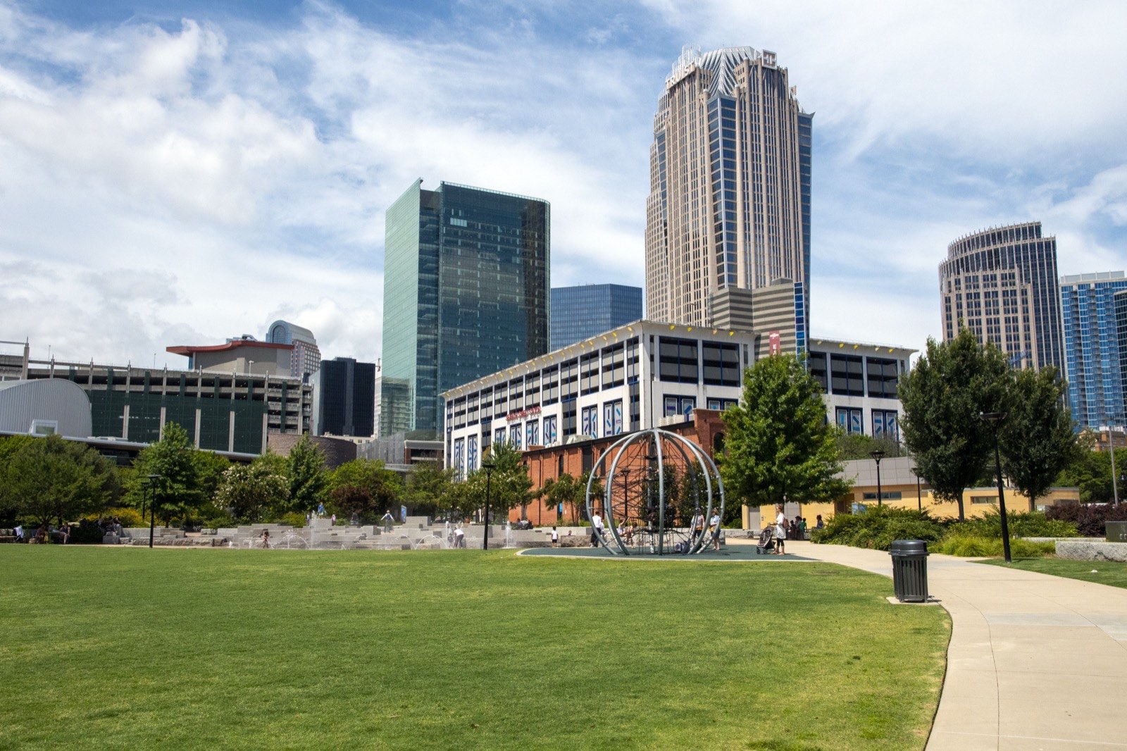 Open lawn and steel sphere sculpture at First Ward Park in Uptown Charlotte, with the Bank of America Corporate Center and other high-rise towers behind