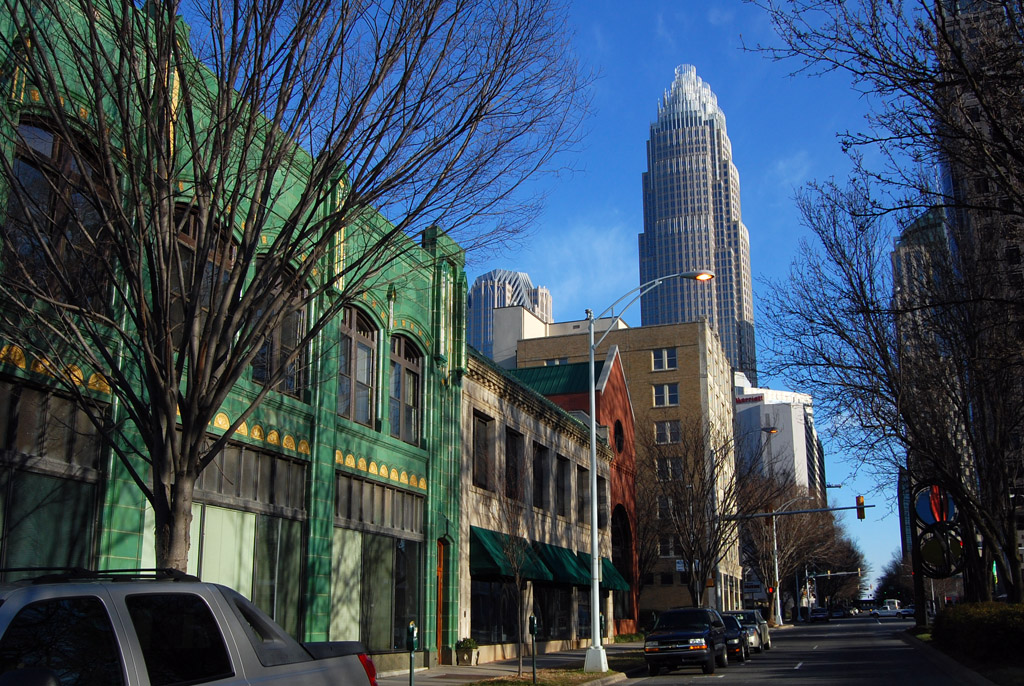 Street-level view of historic green terracotta and brick storefronts on West Trade Street in Uptown Charlotte, with the Bank of America Corporate Center tower rising behind them