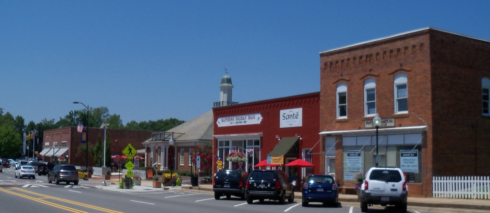 Row of brick storefronts in downtown Matthews, North Carolina, including Matthews Holiday Haus and Sante, with cars parked along Trade Street under a clear blue sky